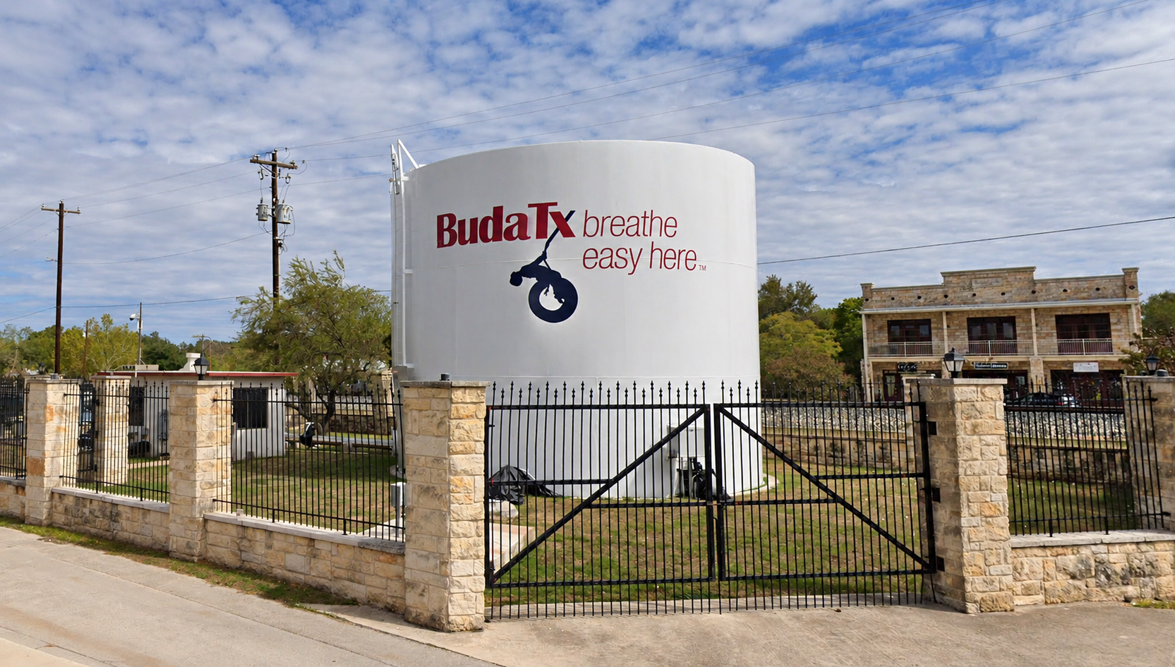 White Buda TX water tank with the words Breathe easy here, behind a stone and iron fence.