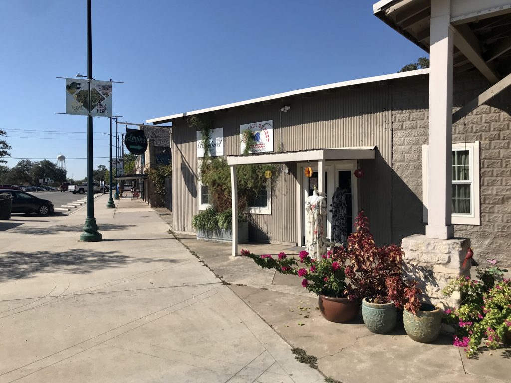 Downtown Buda storefronts line a sunny sidewalk with potted flowers.