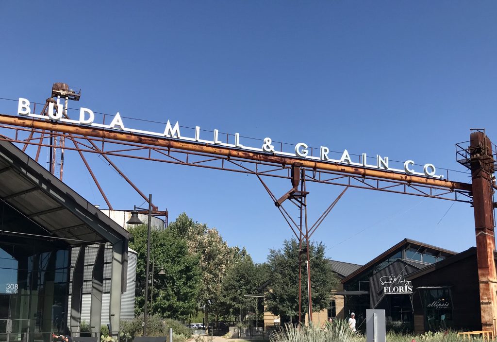 The Buda Mill & Grain Co. sign stretches above shops beneath a blue sky.